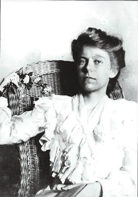 A woman in a lacy dress sits in a woven chair and looks directly at the camera.