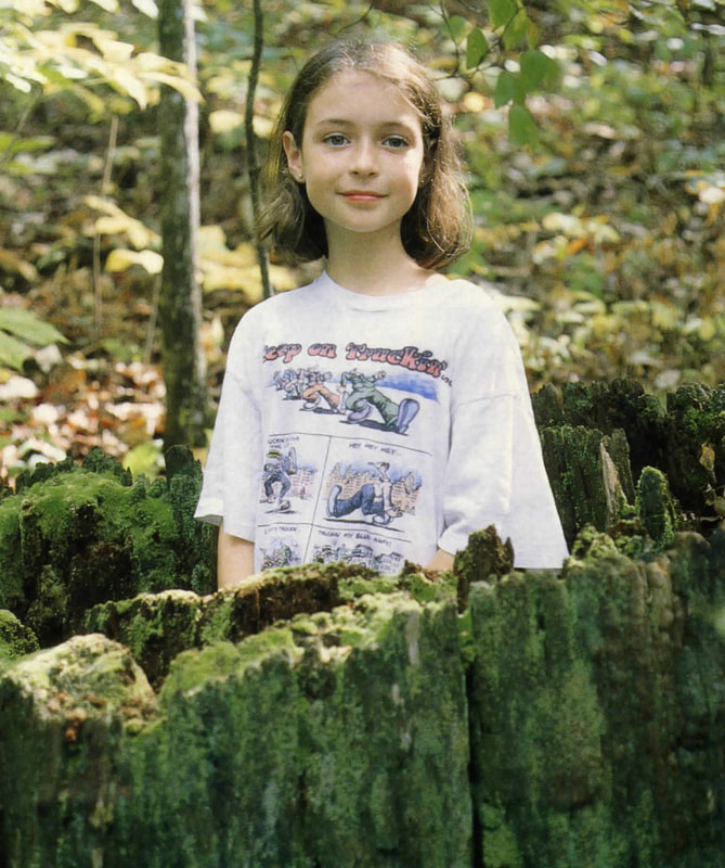 A young girl in a white t-shirt and brown hair poses inside a tree stump on a summer day.