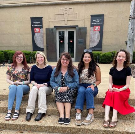 Five women sit on a pebble step in front of a beige building.