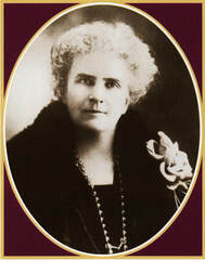 An older lady faces the camera wearing a fur piece, long necklace and with an enormous flower pinned to her left shoulder.