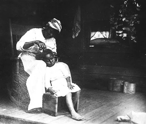 A photograph of an African-American mother sitting in a chair combs her daughter's hair, who is leaning against her knee.