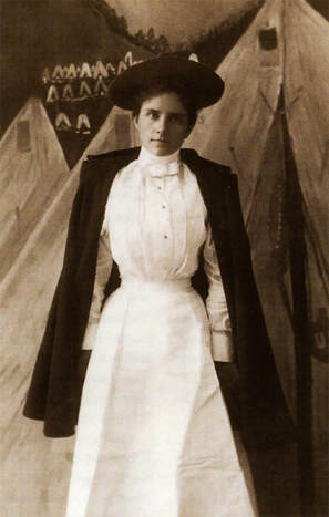 A young woman stands facing the camera and wearing an early 20th century nursing uniform.