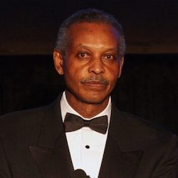 A Black elderly man is shown looking toward the camera. The background is dark, suggesting an evening photo. The man is in formal suit and bowtie, suggesting he is attending a formal event. He has short grey hair, medium-to-dark complexion.